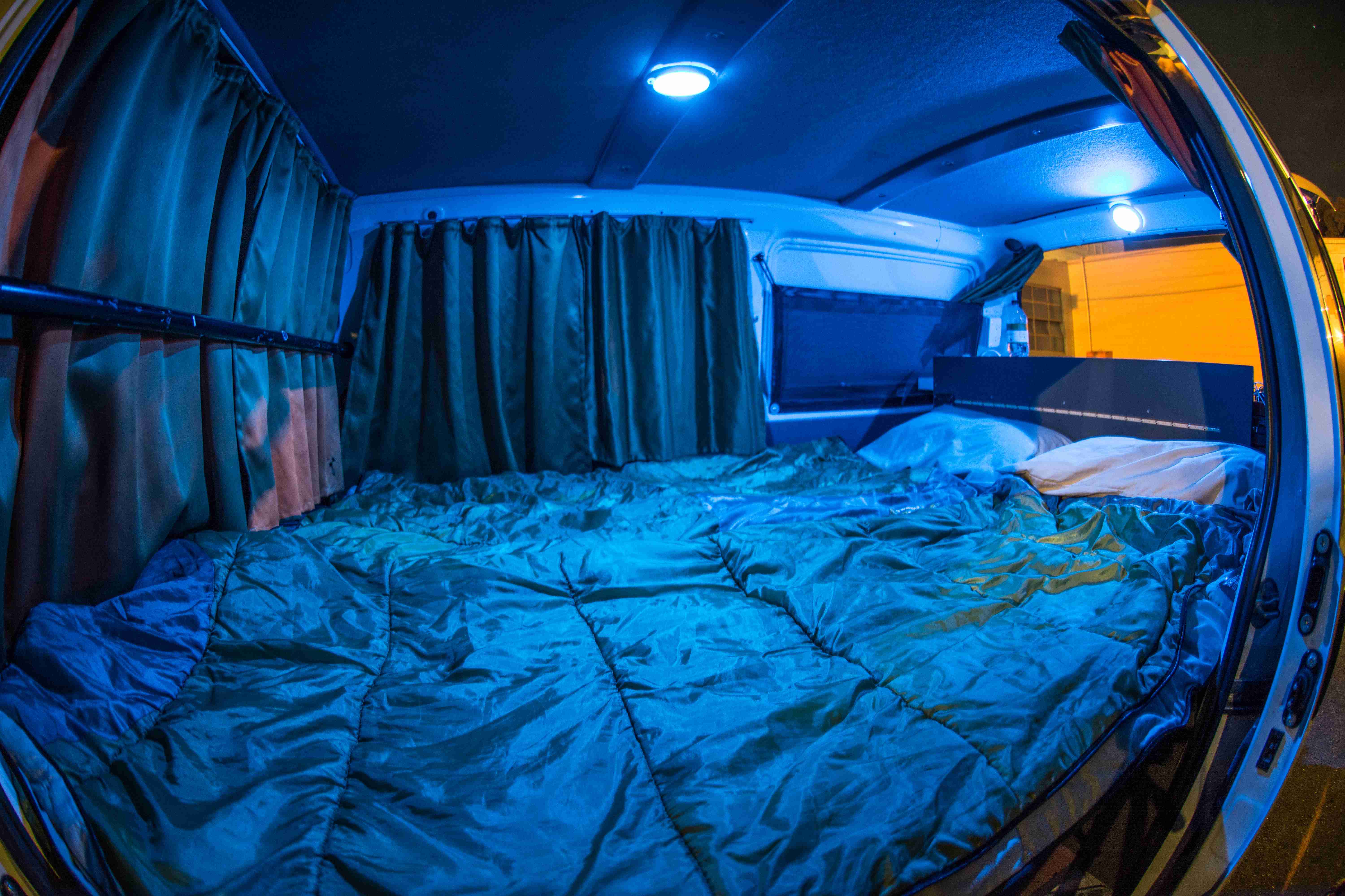Campervan interior at night featuring a bed with a green sleeping bag, dark curtains, and blue ceiling lights.