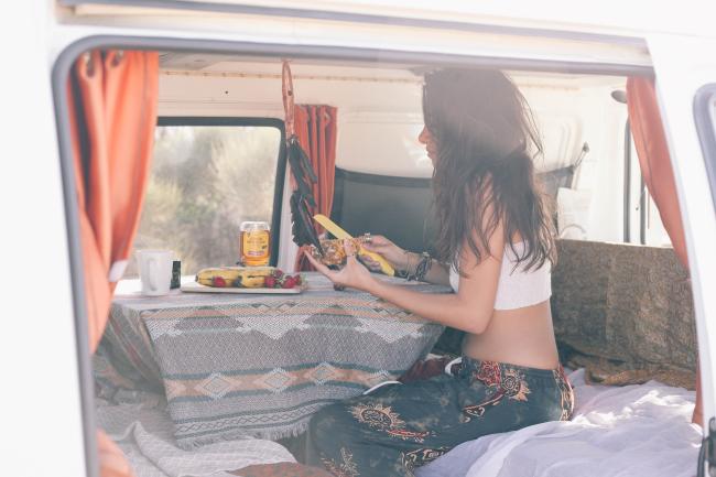A woman sitting inside a campervan prepares food at a table covered in a patterned tablecloth.