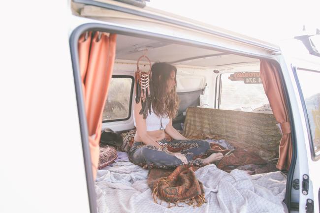 Woman sits cross-legged on a bed inside a white campervan with orange curtains and a dreamcatcher hanging.