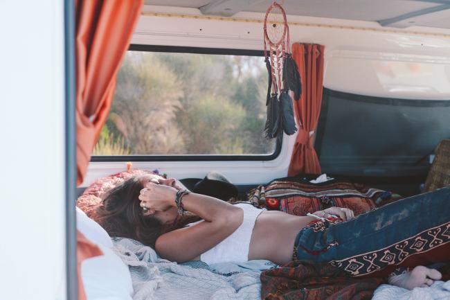 Woman lying down inside a campervan next to a window with orange curtains and a hanging dreamcatcher.