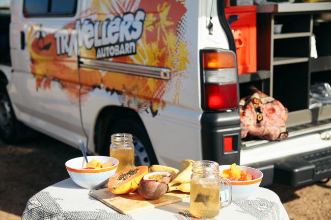 Tropical breakfast of papaya, bananas, and iced tea on a table beside a Travellers Autobarn campervan.