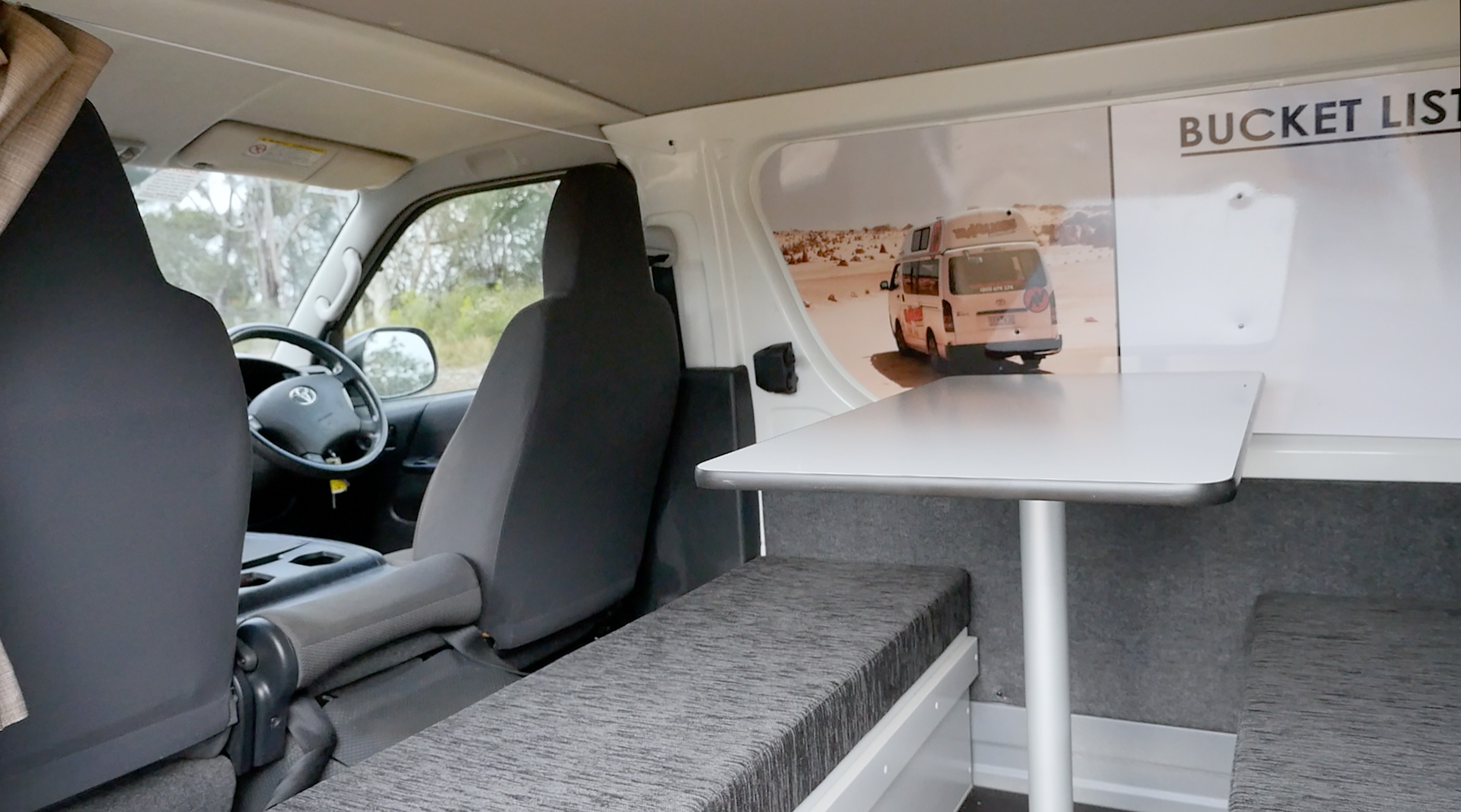Campervan interior showing front grey seats, a grey upholstered bench, and a white dining table with a metal leg.