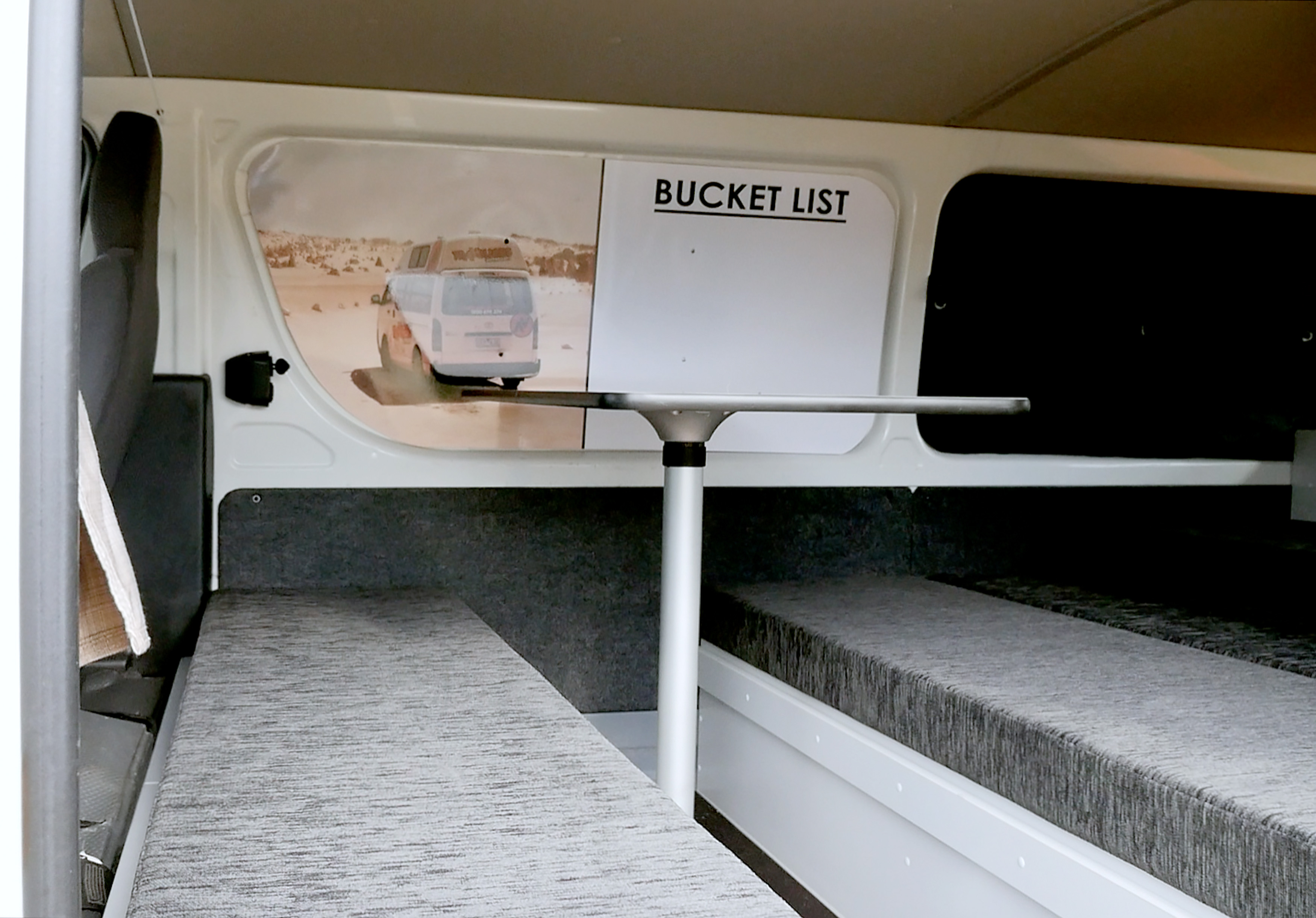 Campervan interior featuring parallel grey cushioned benches, a central pedestal table, and a Bucket List wall decal.