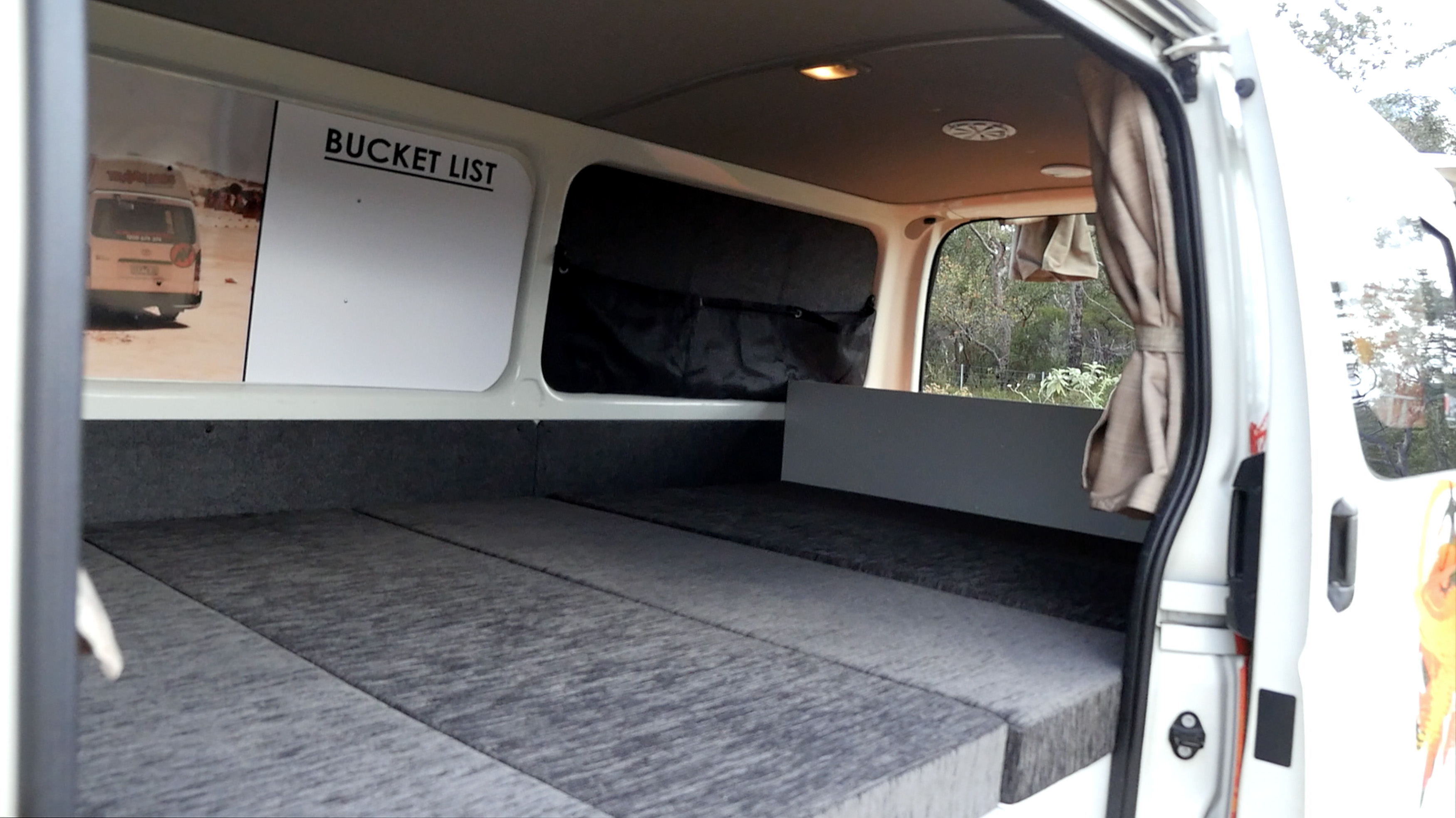 Interior of a white campervan with a grey cushioned bed setup and a wall sign reading 