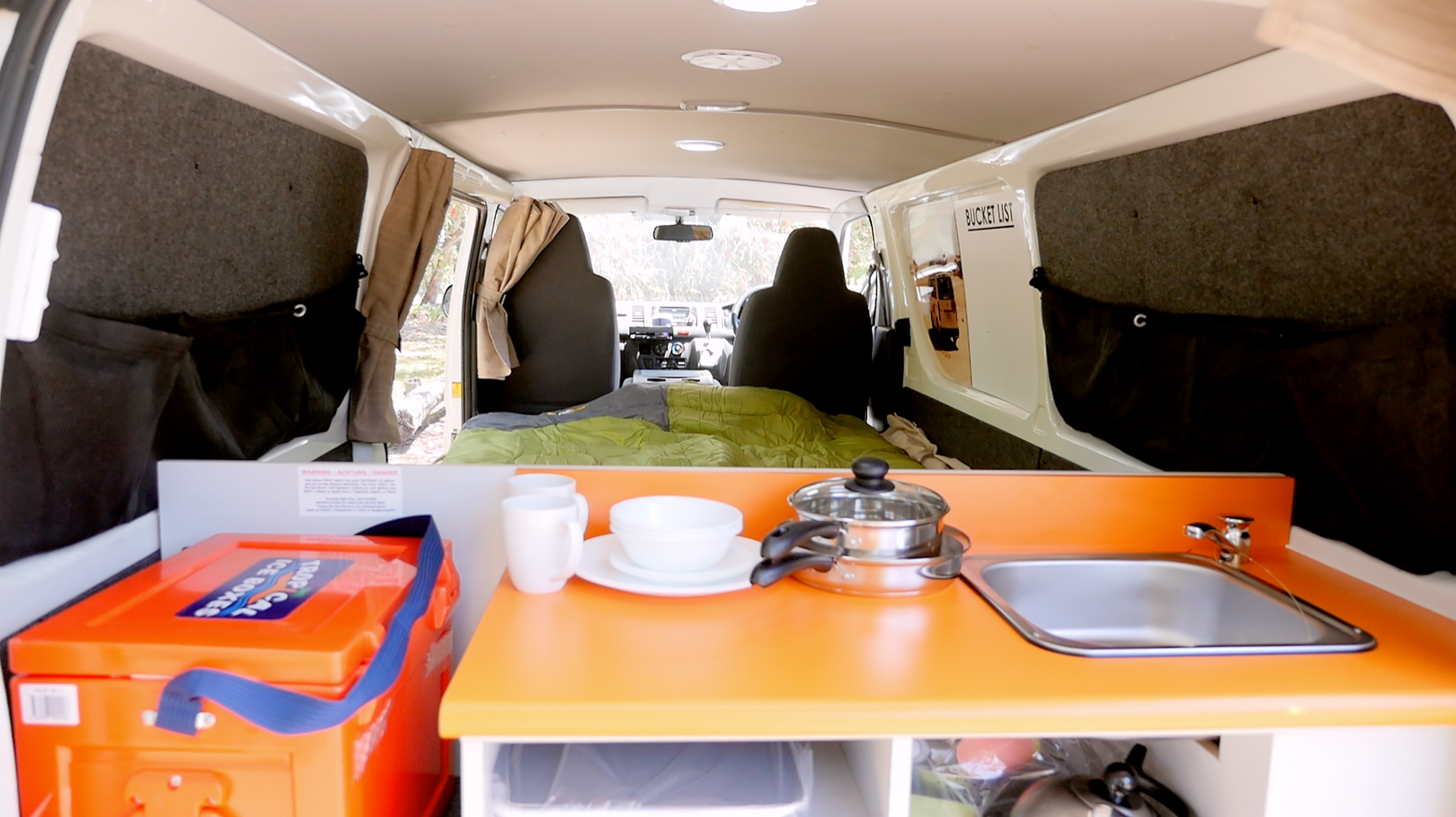 Interior of a campervan showing an orange kitchen counter with a sink, dishes, and a bed in the background.