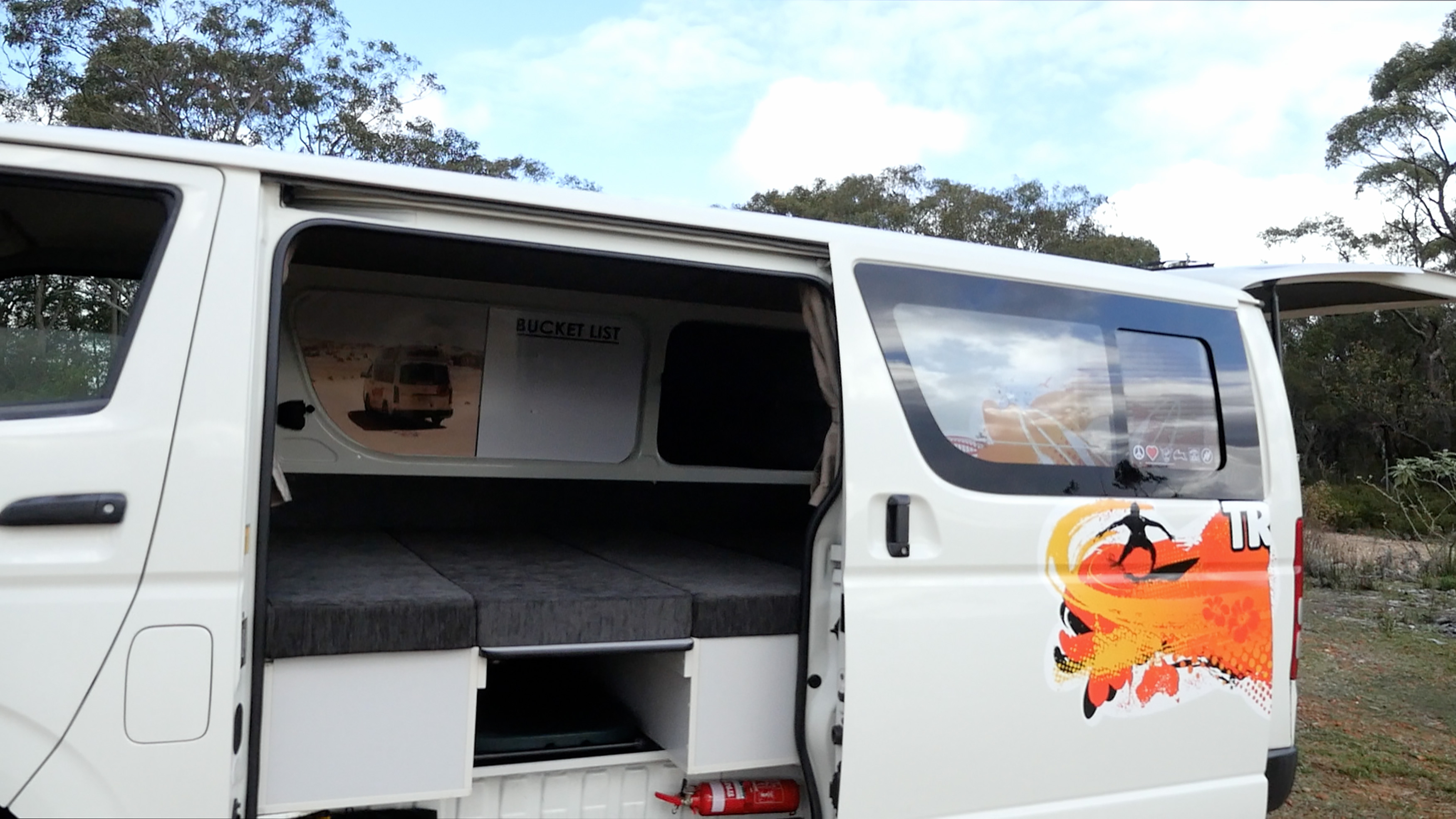White campervan with open sliding door revealing a grey mattress bed, storage underneath, and an exterior surf graphic.