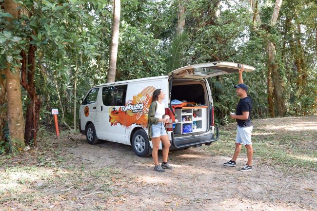 Two travellers stand by the open back of a white campervan parked in a forest clearing.