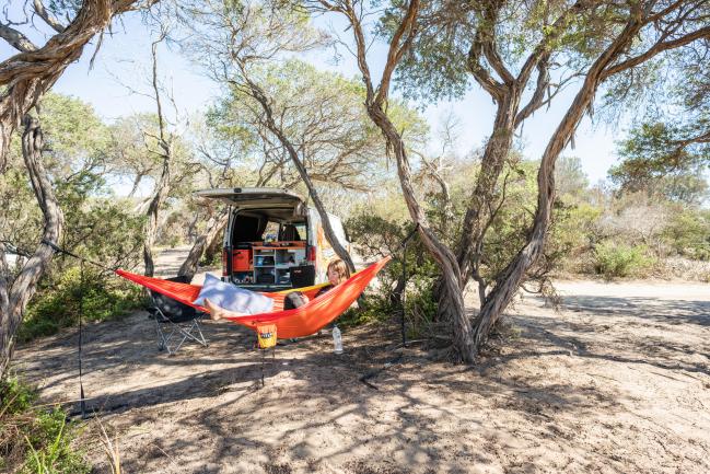 A person relaxes in an orange hammock strung between trees next to a campervan with its back hatch open.