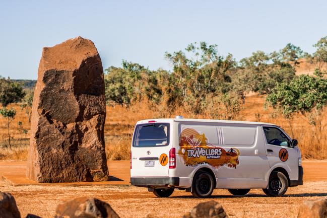 White Travellers Autobarn campervan parked next to a large standing rock in a dry Australian outback landscape.