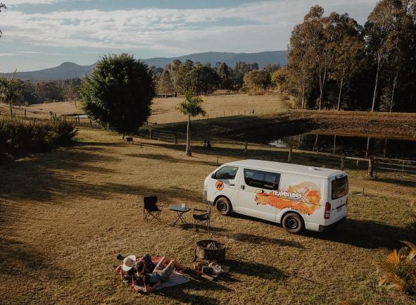 Two people relax on a grassy field near a parked white campervan, with a pond and hills in the background.