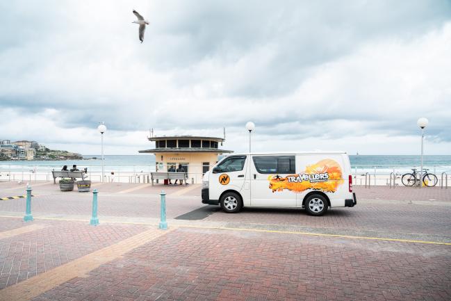 White Travellers campervan parked on a paved beachfront promenade with a lifeguard tower in the background.