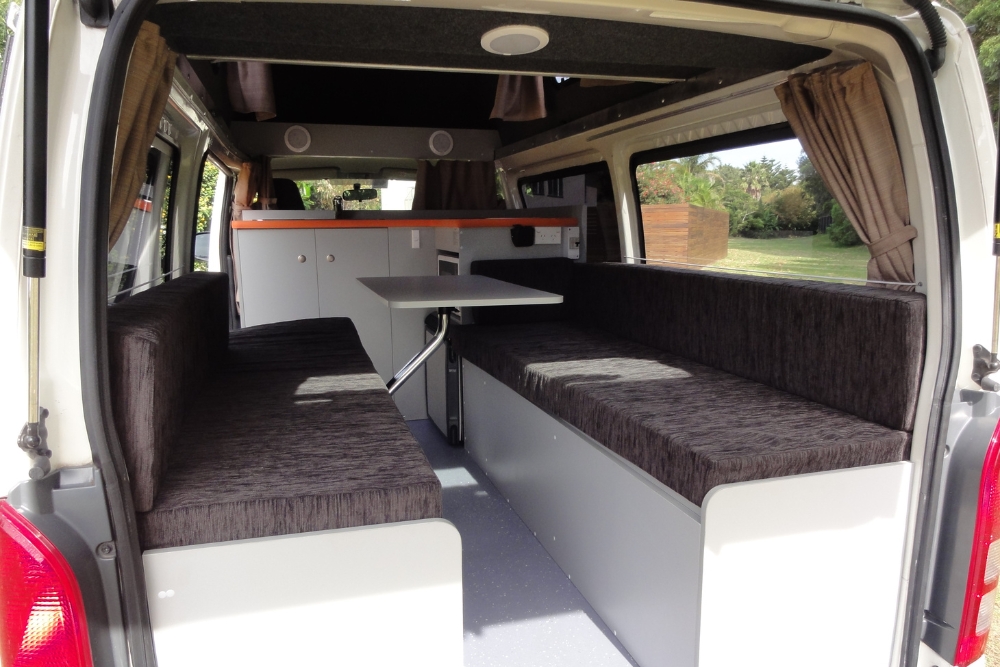 Campervan interior with parallel grey benches, a central table, and kitchenette cabinetry.