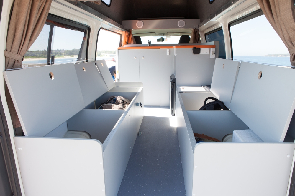 Interior of a campervan with open grey storage benches along a central aisle leading to a front kitchen cabinet.
