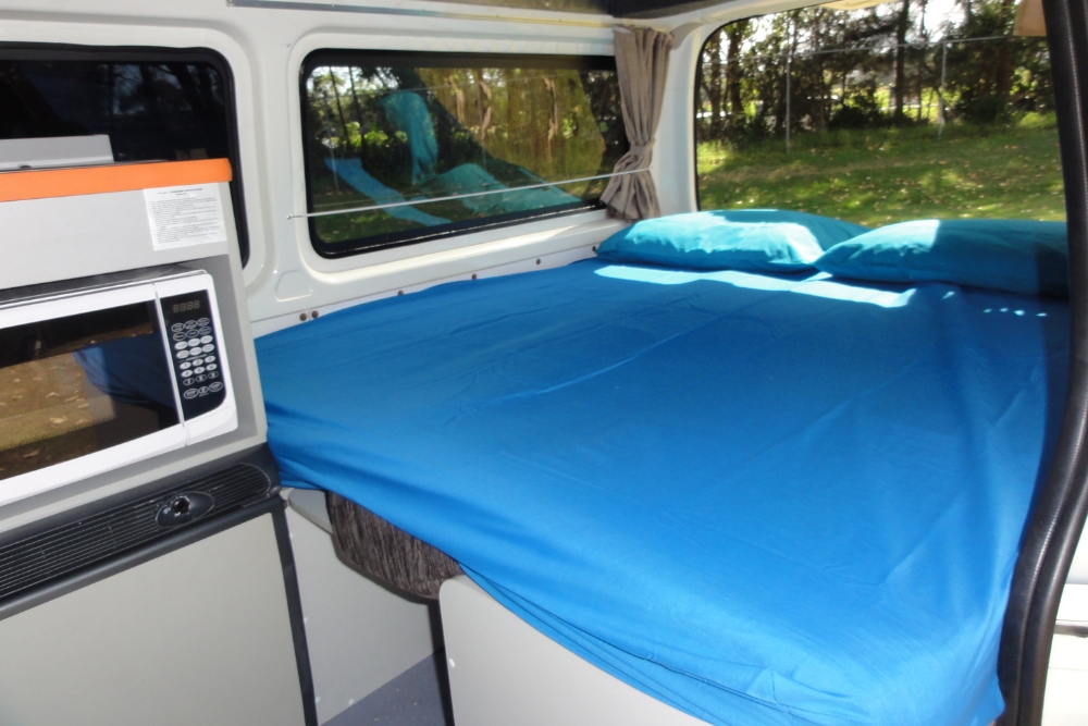 Campervan interior featuring a double bed with blue sheets and pillows next to a kitchenette with a microwave.