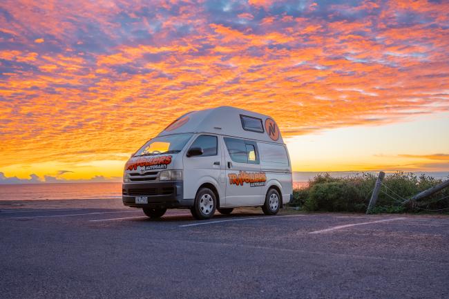 Travellers Autobarn campervan parked in a coastal car park under a bright orange and pink sunset sky.
