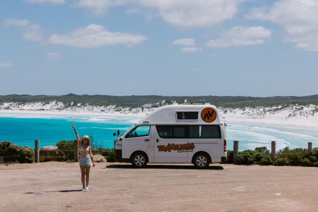 Woman posing next to a white campervan parked overlooking a turquoise beach with white sand.