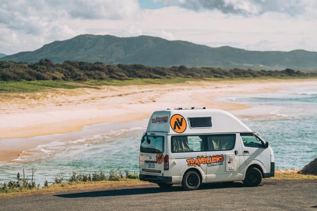 White Travellers Autobarn campervan parked overlooking a sandy beach and turquoise ocean with distant hills.