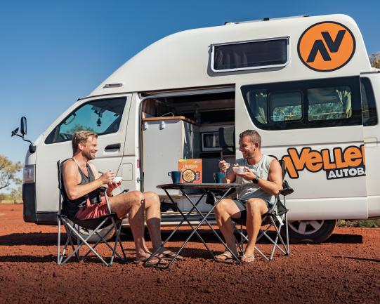 Two men sitting in camp chairs eating meals outside a white campervan parked on red outback dirt.