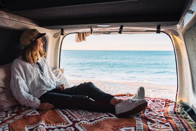 Woman lounging inside a campervan, looking out the open back at a sandy beach and ocean.