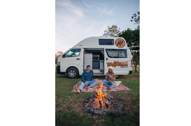 A couple sits on a rug by a campfire in front of a white Travellers Autobarn campervan with its side door open.