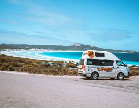 White campervan parked on a coastal road overlooking a turquoise bay and white-sand beach.