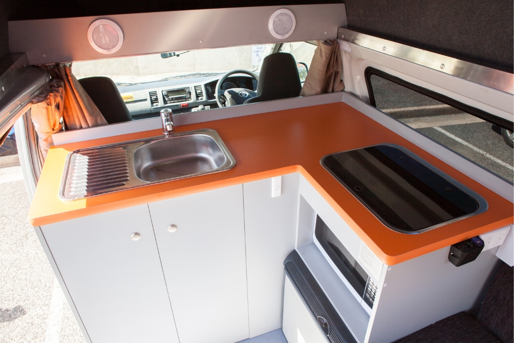 Campervan kitchen featuring an orange benchtop, stainless steel sink, cooktop, microwave, and cabin seats in background.