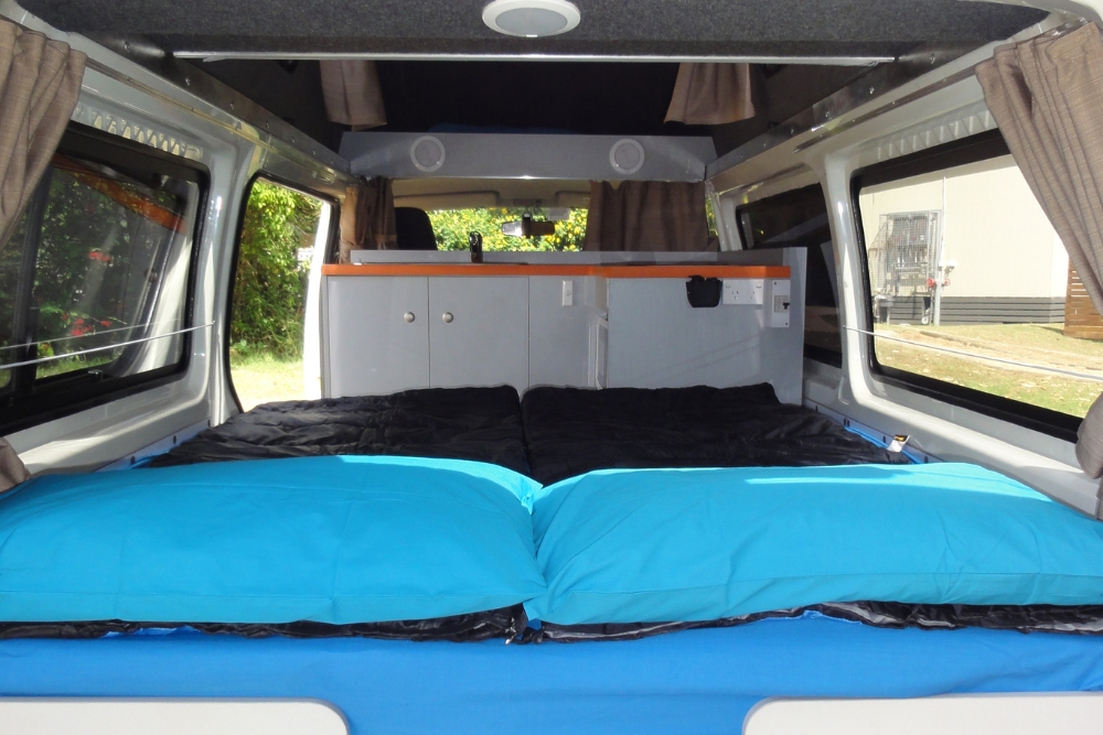 Campervan interior showing a bed with bright blue pillows in front of white kitchen cabinets with orange trim.