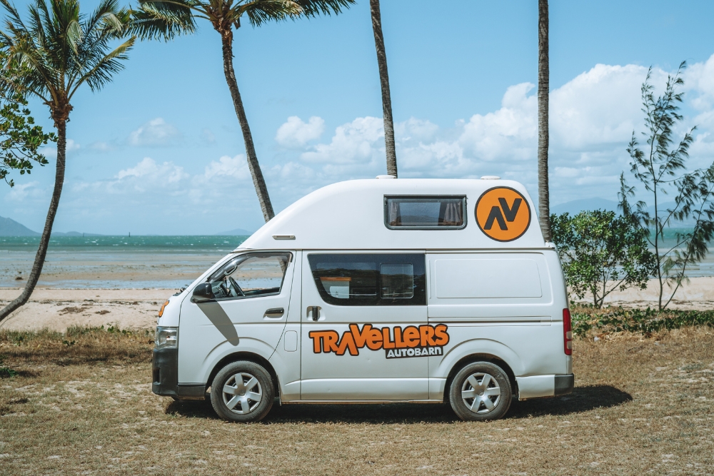 White Travellers Autobarn campervan parked near a sandy beach with palm trees.