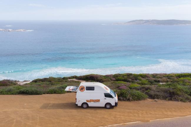 White campervan with its back hatch open parked on a dirt cliff overlooking a turquoise ocean.