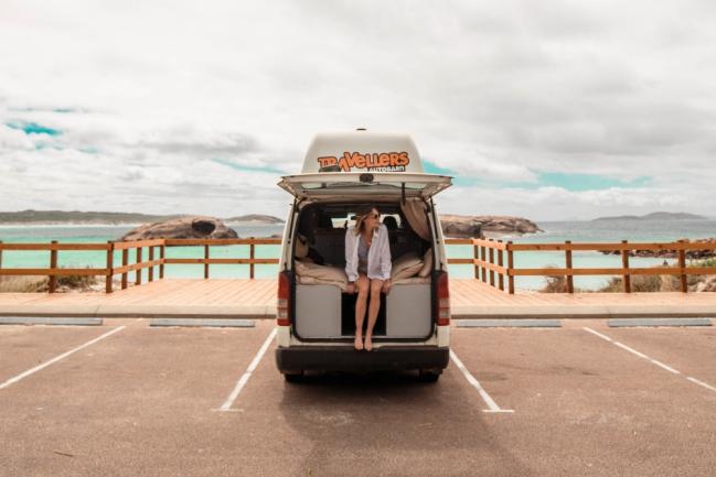 Woman sits in the open back of a campervan parked in a carpark overlooking a turquoise coastal bay.