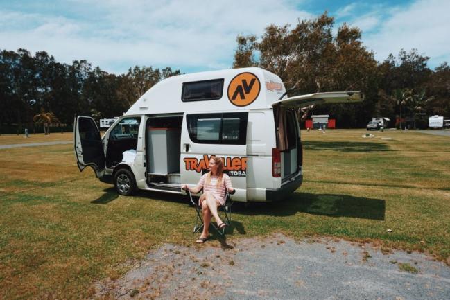 Woman sitting in a folding chair next to a white campervan with its side and rear doors open on a grassy field.