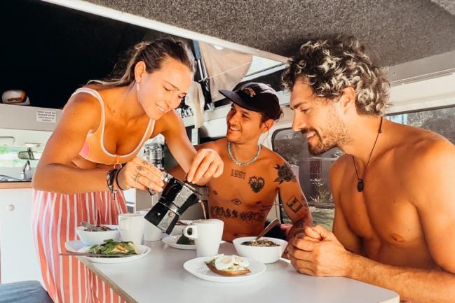 A woman pours coffee from a Moka pot for two men having breakfast inside a campervan.