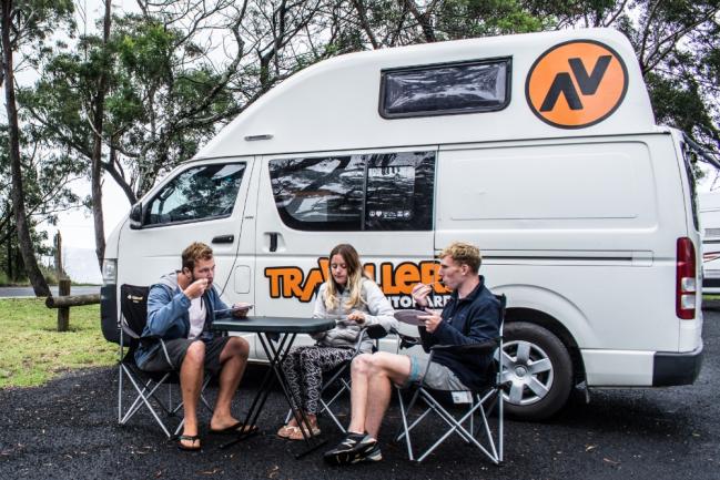 Three people eating at a folding table next to a parked white Travellers campervan.