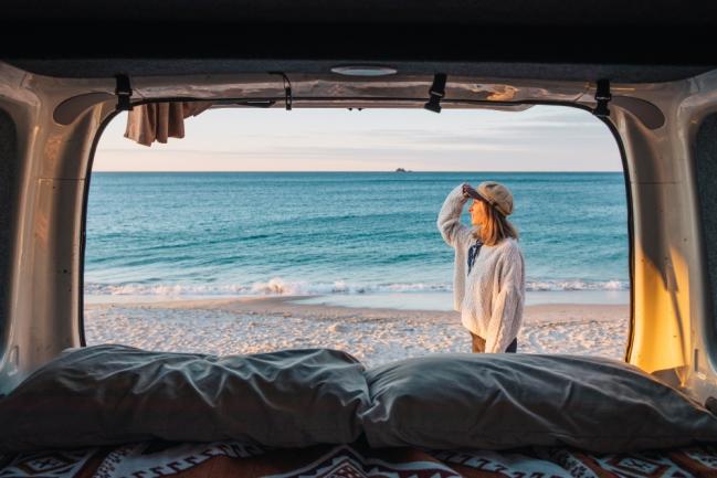 View from the back of a campervan looking out to a woman standing on a sandy beach facing the ocean.