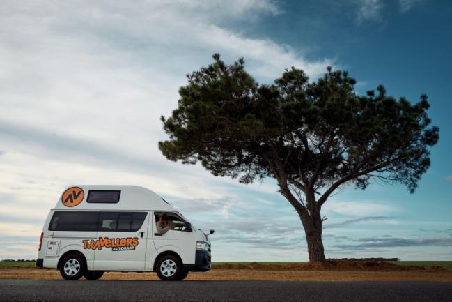 White Travellers Autobarn campervan parked on a roadside next to a large tree under a cloudy sky.
