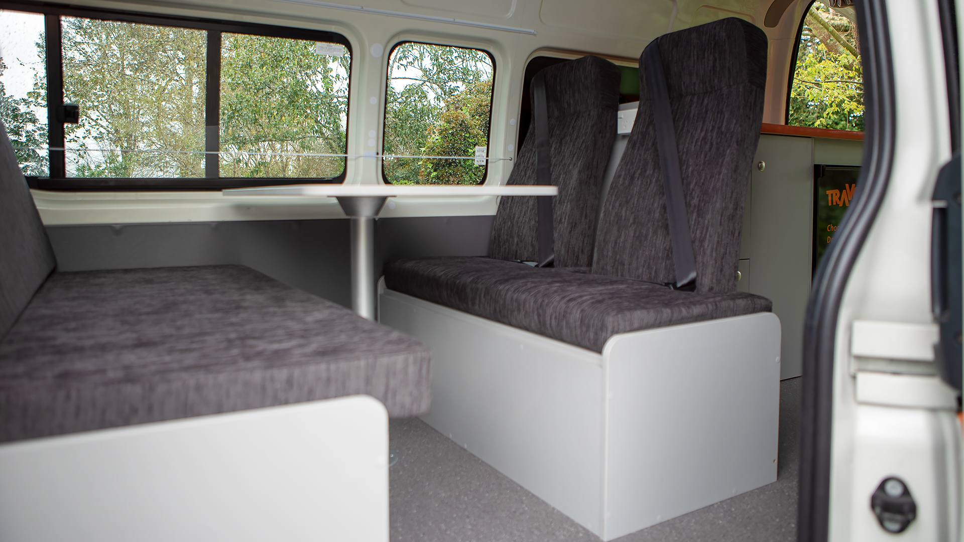 Interior of a campervan with grey upholstered seats, a central table, and high-back passenger seats with seatbelts.