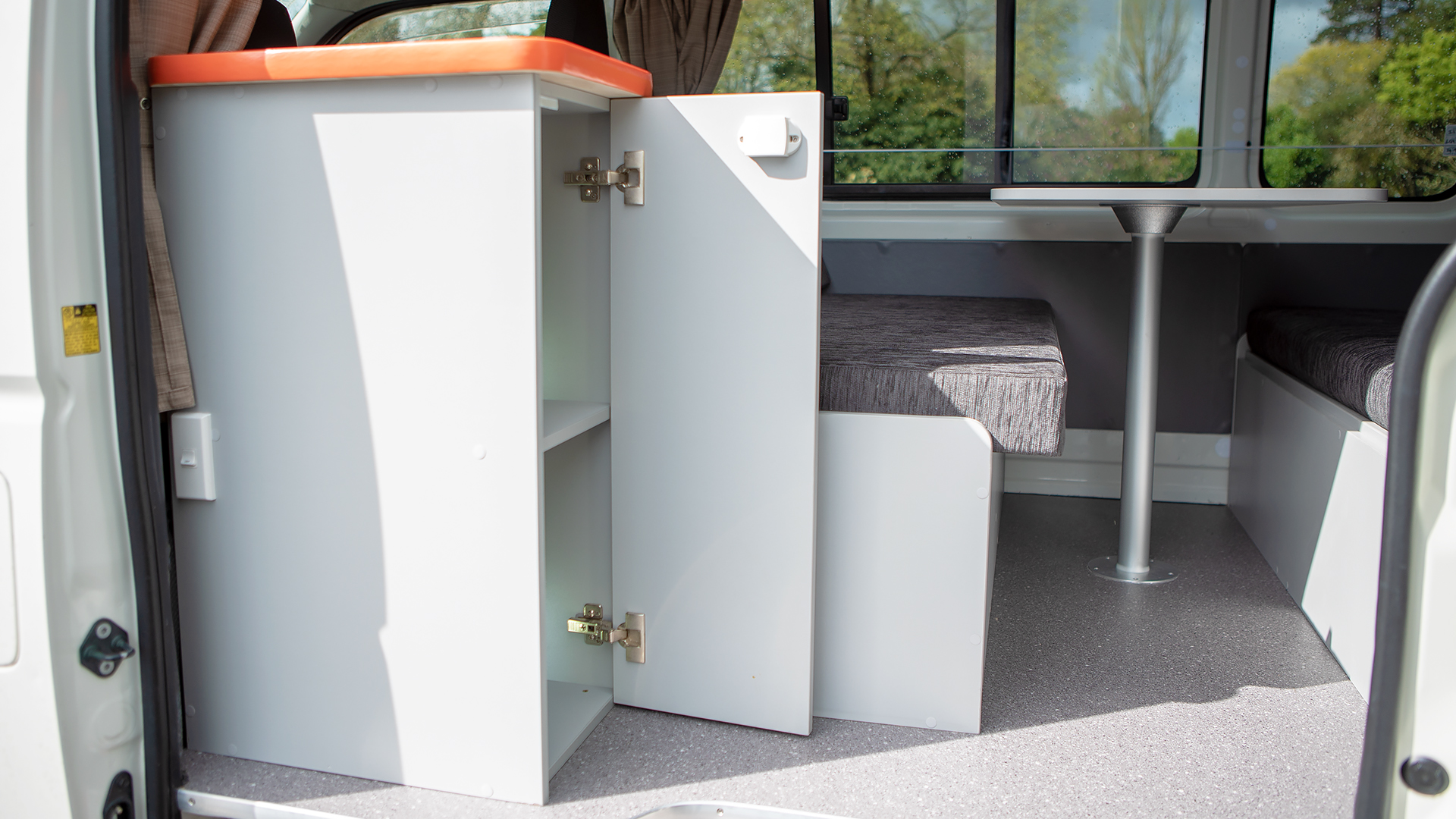 Campervan interior featuring an open white cabinet with an orange top, grey bench seating, and a central table.