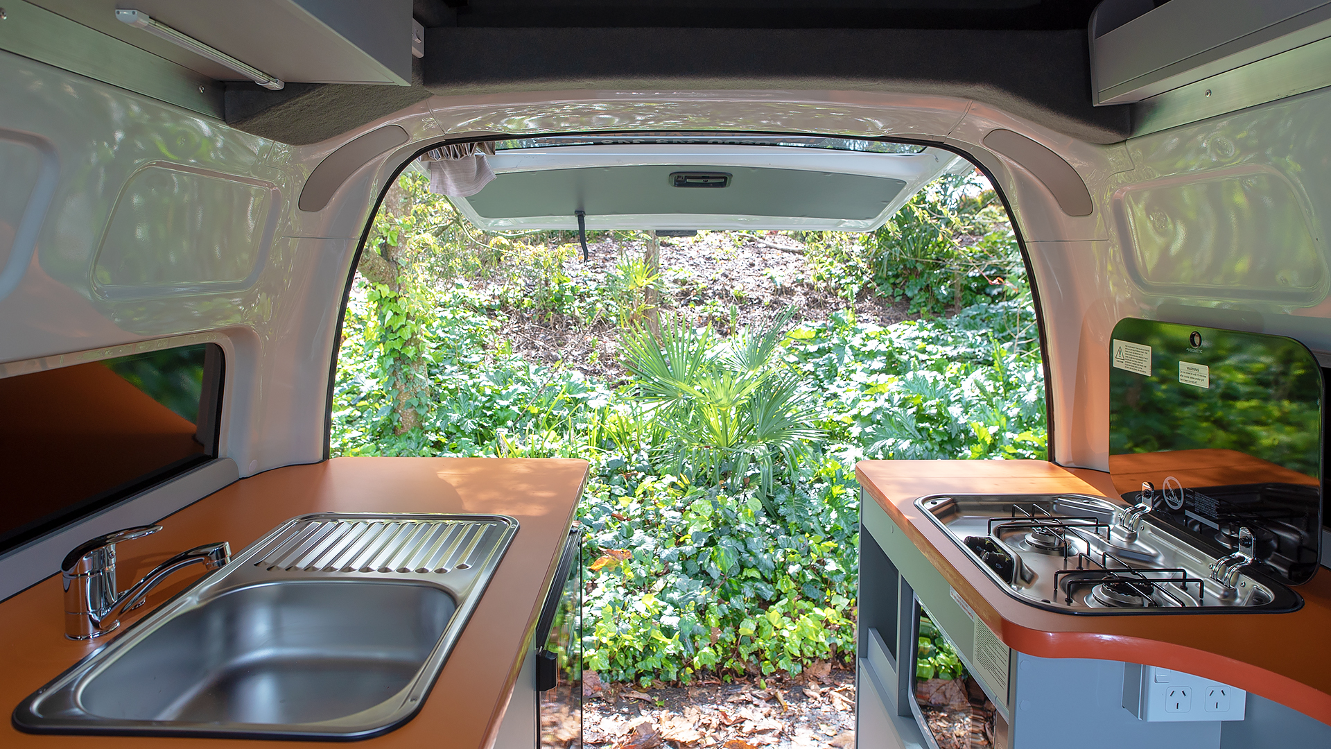Campervan interior looking out the open rear hatch to green foliage, featuring a kitchen sink and a two-burner gas stove.