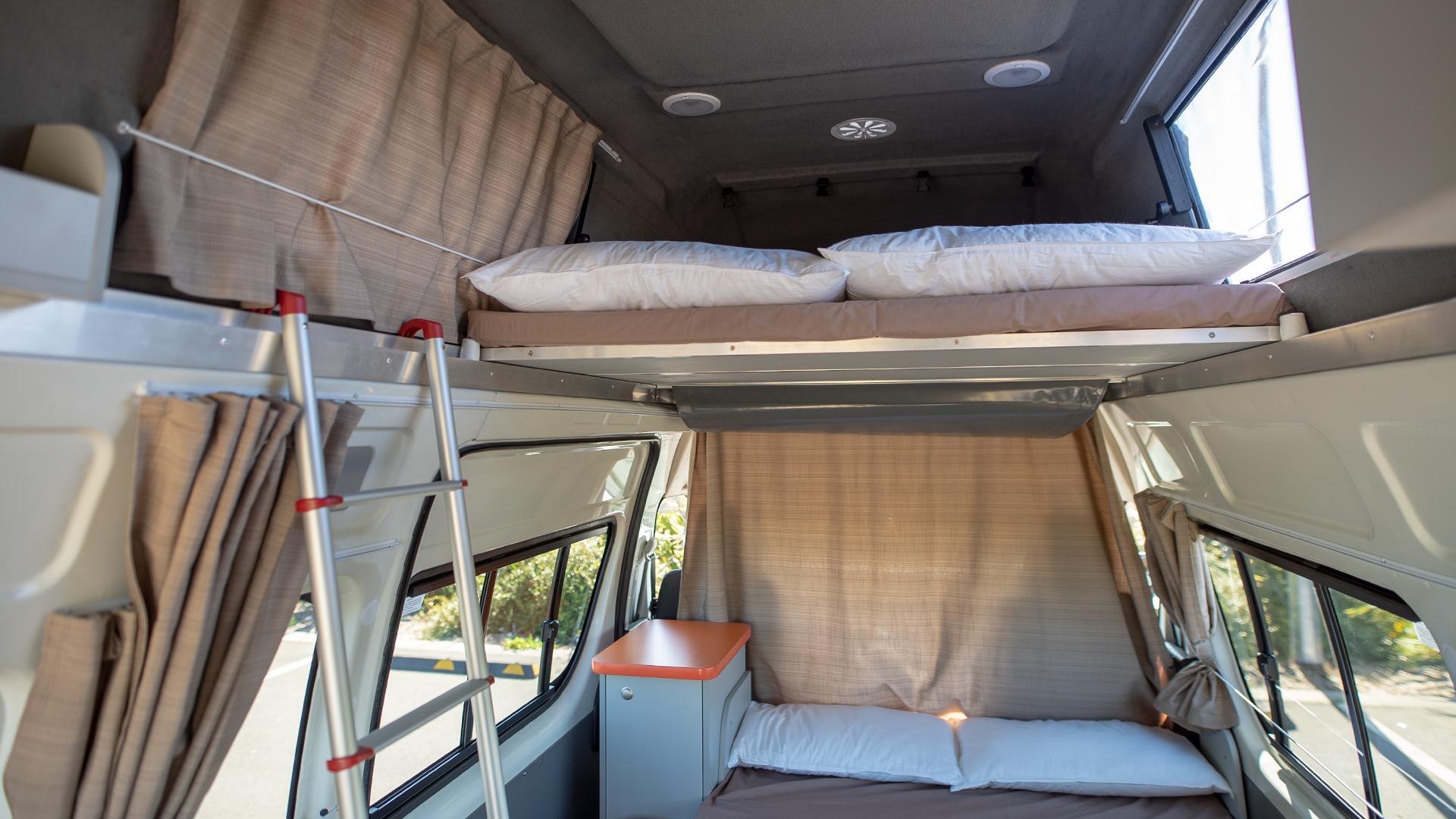 Campervan interior featuring two bunk beds with white pillows, beige curtains, and a metal ladder to the upper bed.