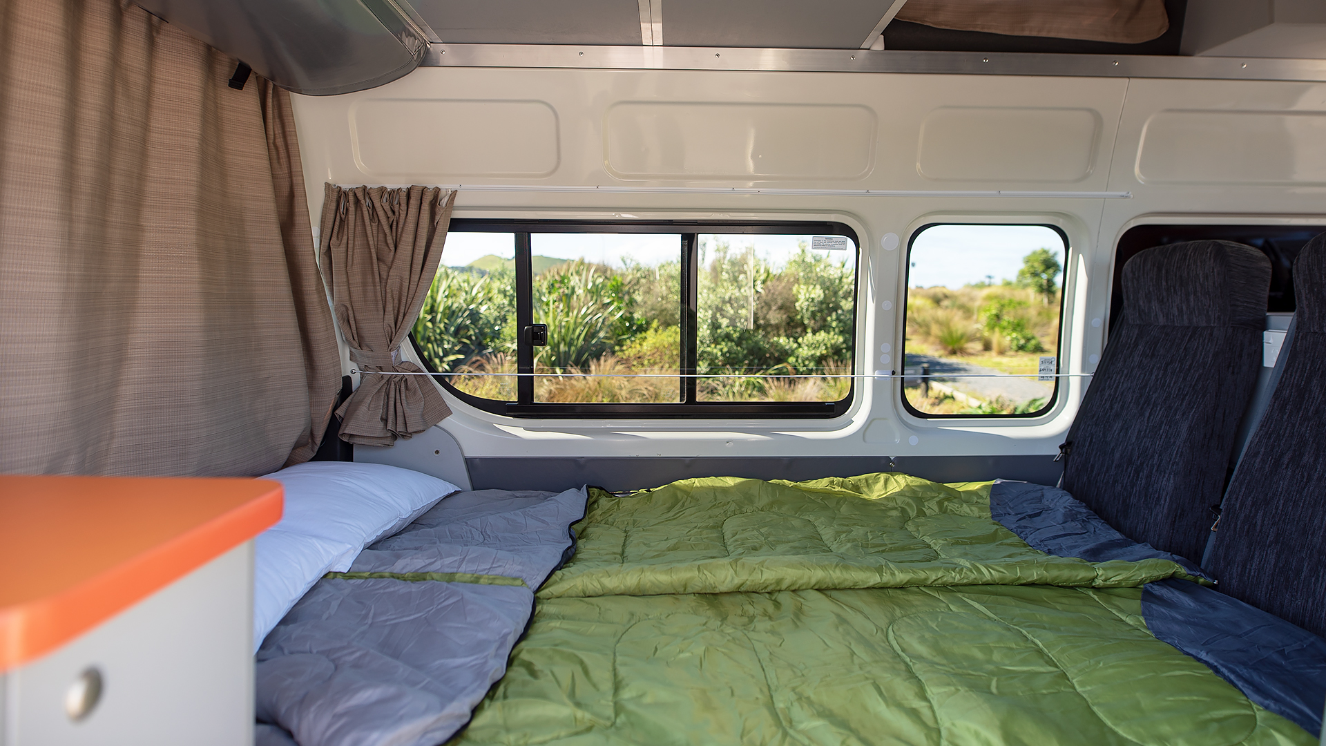 Campervan interior featuring a bed with a green sleeping bag, white pillow, and windows overlooking foliage.