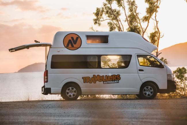White Travellers Autobarn campervan with open tailgate parked on a coastal road at sunset.