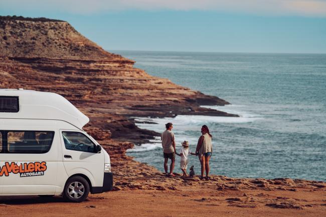 White campervan parked on a rugged coastal cliff with a family standing near the edge looking at the ocean.