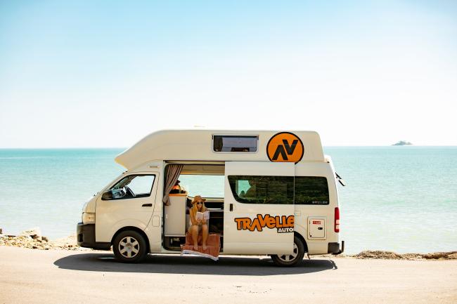 Woman reading a book in the open doorway of a white campervan parked beside a calm blue sea.