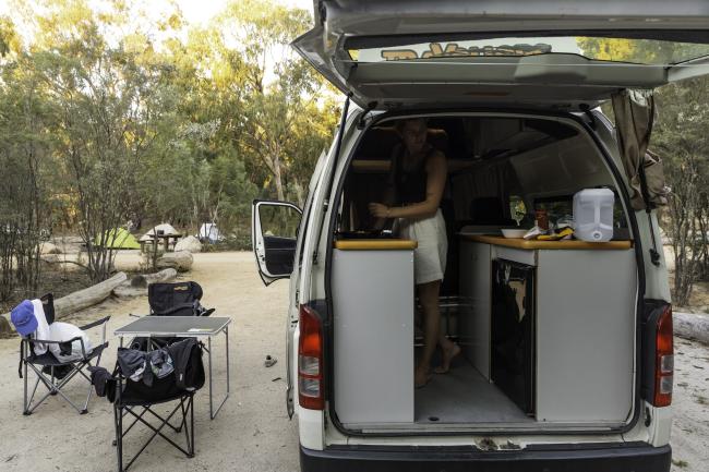 Woman stands inside a white campervan with an open rear hatch at a campsite with outdoor chairs and a table.
