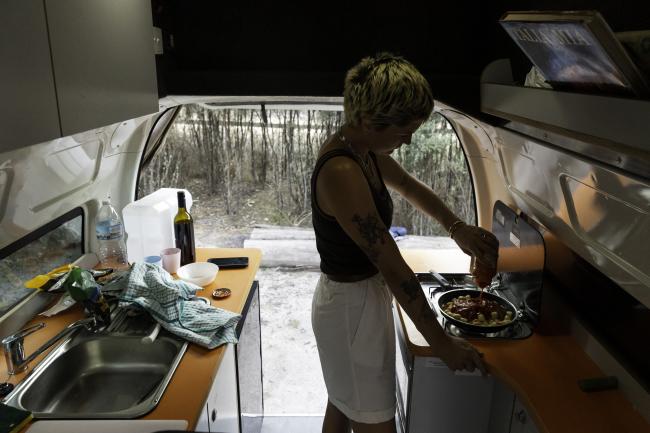 A person cooks inside a campervan, pouring sauce into a pan on a stove with open back doors showing a forest.