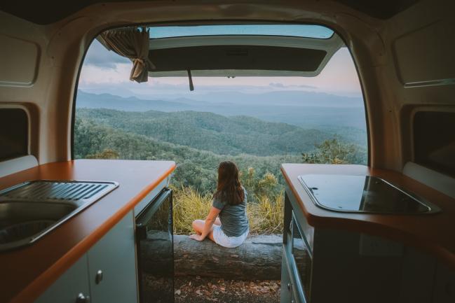 View from inside a campervan looking past countertops to a woman on a log overlooking forested mountains.