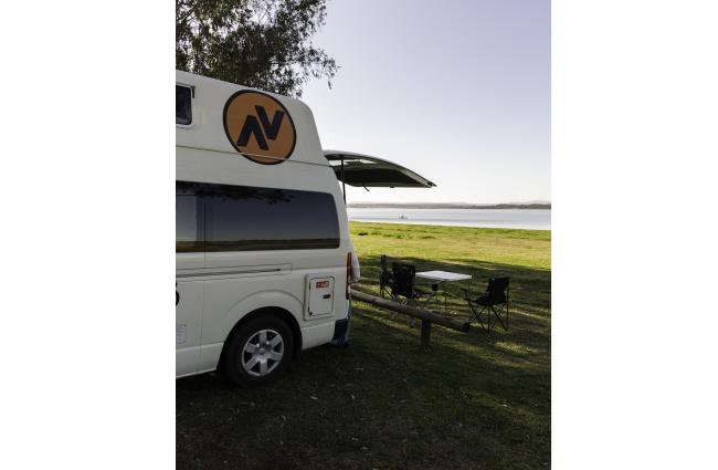 White campervan parked by a lake, with camping chairs and a table set up on the grass near the open rear hatch.