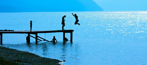 silhouettes of kids who jump off dock on the lake blue toned